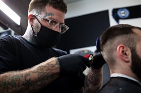 A barber with tattoos on his arms and forehead is wearing a mask and gloves while using clippers to cut a customer's hair. The setting appears to be a barber shop, with a dark background and bright lighting focused on the activity.