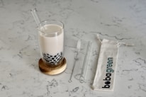 A glass of milk tea with black tapioca pearls sits on a wooden coaster. Beside it, a transparent glass straw and a cleaning brush are placed on a light-colored marble surface. A fabric bag with 'bobagreen' printed on it lies flat.