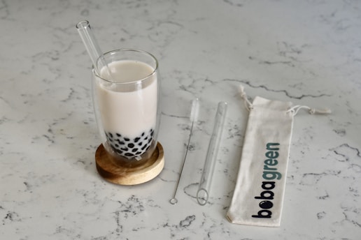 A glass of milk tea with black tapioca pearls sits on a wooden coaster. Beside it, a transparent glass straw and a cleaning brush are placed on a light-colored marble surface. A fabric bag with 'bobagreen' printed on it lies flat.