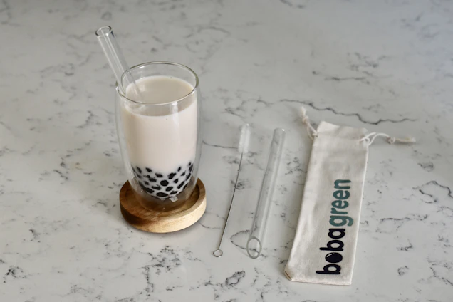 A glass of milk tea with black tapioca pearls sits on a wooden coaster. Beside it, a transparent glass straw and a cleaning brush are placed on a light-colored marble surface. A fabric bag with 'bobagreen' printed on it lies flat.