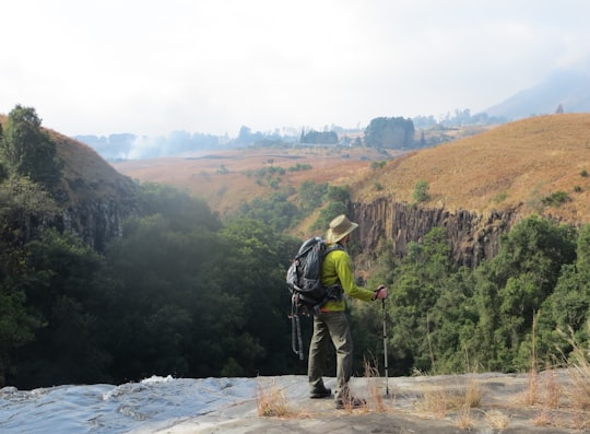 man in green jacket and brown pants standing on rock near river during daytime in Drakensberg South Africa