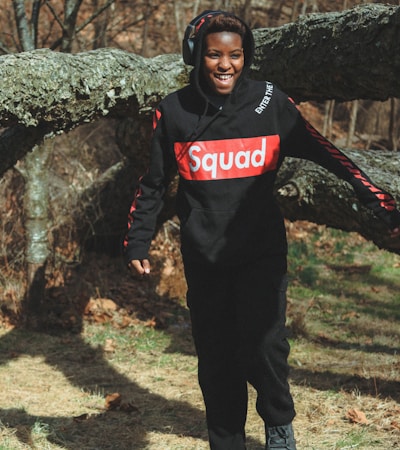 A person wearing a black hoodie with 'Squad' written in red is smiling while walking outdoors. They are wearing black pants and headphones. The background features a large, fallen tree and dry, brown foliage, indicative of autumn.
