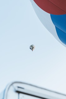 A large hot air balloon is partially visible in the foreground, with vibrant red, white, and blue sections. In the distance, a second hot air balloon floats against a clear blue sky. The image creates a sense of vastness and height.