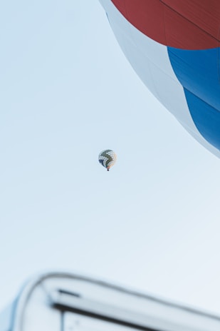A large hot air balloon is partially visible in the foreground, with vibrant red, white, and blue sections. In the distance, a second hot air balloon floats against a clear blue sky. The image creates a sense of vastness and height.