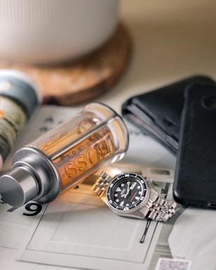 A watch with a metallic strap lies on a flat surface alongside a bottle of Hugo Boss cologne. Nearby is a black leather wallet and a dark smartphone. The setting appears organized, suggesting a focus on personal accessories.