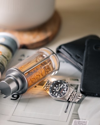 A watch with a metallic strap lies on a flat surface alongside a bottle of Hugo Boss cologne. Nearby is a black leather wallet and a dark smartphone. The setting appears organized, suggesting a focus on personal accessories.