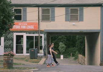 A building with a 'Store for Lease' sign hangs over a small outdoor path leading to a wooded area. Two people, one in a black shirt and pants and the other in a dark top and light pants, walk together in the foreground. A barrel with flowers and an air conditioning unit are visible near the entrance.