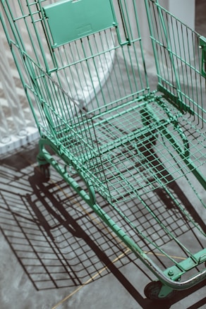A green shopping cart is positioned on a concrete surface, casting a complex shadow. The lighting creates a sharp contrast between the cart and the ground, highlighting the cart's metal grid structure.