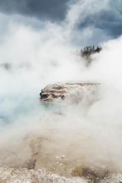 A geothermal landscape with steam rising over rocky terrain under a cloudy sky.