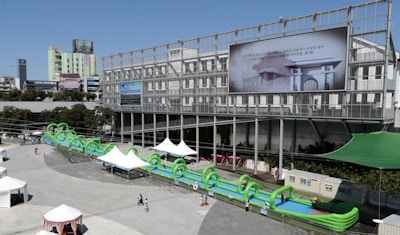 A large inflatable water slide with green arches is set up in an urban outdoor plaza. The slide extends in front of a modern, industrial-style building with a large billboard displaying an image and text in a non-English language. Several white tents are scattered around the area, providing shade for visitors. People are walking around the plaza, enjoying the sunny weather.