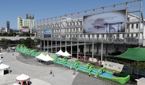 A large inflatable water slide with green arches is set up in an urban outdoor plaza. The slide extends in front of a modern, industrial-style building with a large billboard displaying an image and text in a non-English language. Several white tents are scattered around the area, providing shade for visitors. People are walking around the plaza, enjoying the sunny weather.