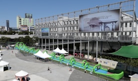A large inflatable water slide with green arches is set up in an urban outdoor plaza. The slide extends in front of a modern, industrial-style building with a large billboard displaying an image and text in a non-English language. Several white tents are scattered around the area, providing shade for visitors. People are walking around the plaza, enjoying the sunny weather.