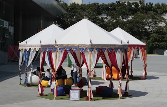 A series of white tents are set up outdoors with vibrant red, blue, and white curtains. Inside, people are seated on colorful bean bags on a patch of artificial grass. The surroundings include trees and a modern building.