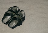 Two green sandals with black buckles are placed on a sandy surface, casting a small shadow.