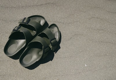 Two green sandals with black buckles are placed on a sandy surface, casting a small shadow.