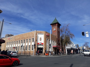 A street corner featuring a historic brick building with a prominent clock tower and a green roof. The building has a series of tall windows and is branded with a bank logo. There are pedestrians walking on the sidewalk and cars parked and driving on the road. The sky is clear and blue, suggesting a sunny day.