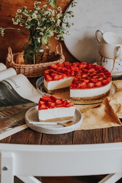 A strawberry-topped cheesecake with a slice placed on a small plate beside it. The setting includes a rustic wooden table, a wicker basket, a glass vase with small white flowers, and stacked ceramic cups on a stand. A magazine or book is casually opened near the cake.