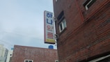A brick building with a visible side featuring a sign with Korean characters and numbers. The building has windows and is adjacent to power lines. The sky is overcast.