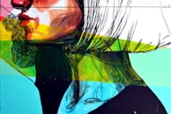 A vibrant street art mural featuring a stylized side profile of a face with bold graphic lines and vivid colors. The artwork combines brush strokes and geometric shapes with a palette of reds, greens, blues, and yellows, creating a dynamic and modern aesthetic.
