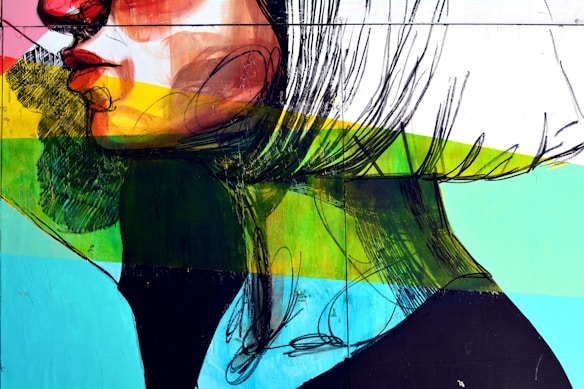 A vibrant street art mural featuring a stylized side profile of a face with bold graphic lines and vivid colors. The artwork combines brush strokes and geometric shapes with a palette of reds, greens, blues, and yellows, creating a dynamic and modern aesthetic.