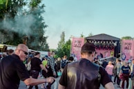 A crowd of people gathered at an outdoor music festival, Pori Jazz 2019, with a stage set up in the background featuring large pink banners. The audience includes individuals engaged in conversation and observing the performance, with some wearing sunglasses and hats. Trees and smoke create a lively atmosphere.