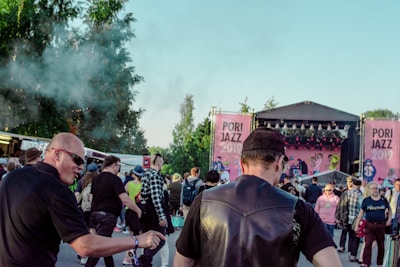 A crowd of people gathered at an outdoor music festival, Pori Jazz 2019, with a stage set up in the background featuring large pink banners. The audience includes individuals engaged in conversation and observing the performance, with some wearing sunglasses and hats. Trees and smoke create a lively atmosphere.