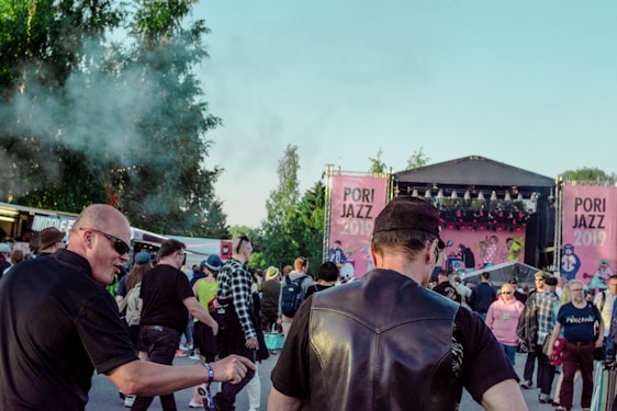 A crowd of people gathered at an outdoor music festival, Pori Jazz 2019, with a stage set up in the background featuring large pink banners. The audience includes individuals engaged in conversation and observing the performance, with some wearing sunglasses and hats. Trees and smoke create a lively atmosphere.