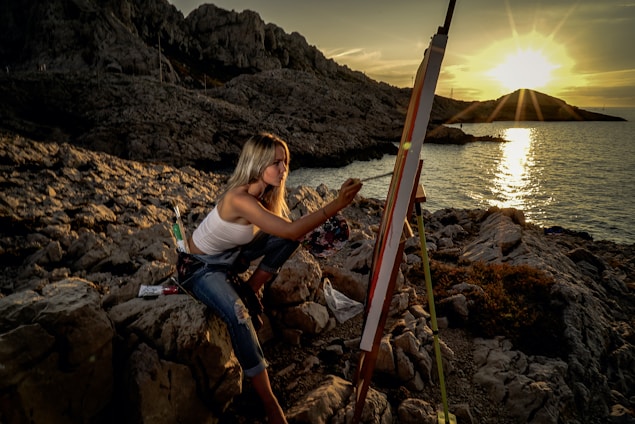 Rachael Townsend standing thoughtfully on a rocky shoreline, with her easel and canvas beside her, waves crashing in the background.