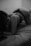 A person is lying on a bed, wearing dark-colored lingerie. The scene is in black and white, creating a dramatic contrast between light and shadow. The setting appears to be a dimly lit room with minimal decor.