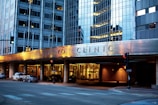 The image depicts the exterior of a modern building with reflective glass windows, displaying the name Mayo Clinic prominently. The building features urban architectural design elements and is well-lit in the evening, creating a warm atmosphere. Several cars are parked along the street, and there are traffic lights at the intersection.