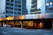 The image depicts the exterior of a modern building with reflective glass windows, displaying the name Mayo Clinic prominently. The building features urban architectural design elements and is well-lit in the evening, creating a warm atmosphere. Several cars are parked along the street, and there are traffic lights at the intersection.