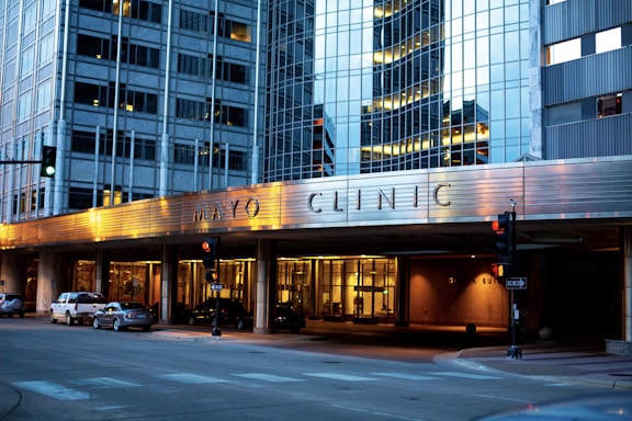 The image depicts the exterior of a modern building with reflective glass windows, displaying the name Mayo Clinic prominently. The building features urban architectural design elements and is well-lit in the evening, creating a warm atmosphere. Several cars are parked along the street, and there are traffic lights at the intersection.