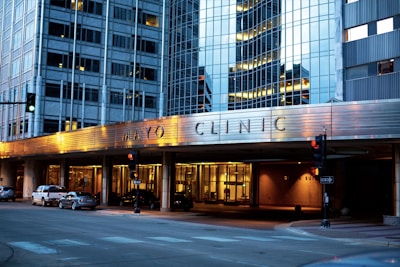 The image depicts the exterior of a modern building with reflective glass windows, displaying the name Mayo Clinic prominently. The building features urban architectural design elements and is well-lit in the evening, creating a warm atmosphere. Several cars are parked along the street, and there are traffic lights at the intersection.