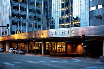 The image depicts the exterior of a modern building with reflective glass windows, displaying the name Mayo Clinic prominently. The building features urban architectural design elements and is well-lit in the evening, creating a warm atmosphere. Several cars are parked along the street, and there are traffic lights at the intersection.