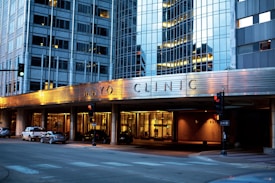 The image depicts the exterior of a modern building with reflective glass windows, displaying the name Mayo Clinic prominently. The building features urban architectural design elements and is well-lit in the evening, creating a warm atmosphere. Several cars are parked along the street, and there are traffic lights at the intersection.