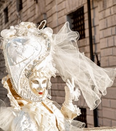 An elaborate Venetian costume features an intricately designed mask with golden embellishments and a large headdress adorned with pearls and lace, topped with delicate veils. The background consists of an old stone building with small barred windows.