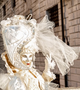 An elaborate Venetian costume features an intricately designed mask with golden embellishments and a large headdress adorned with pearls and lace, topped with delicate veils. The background consists of an old stone building with small barred windows.