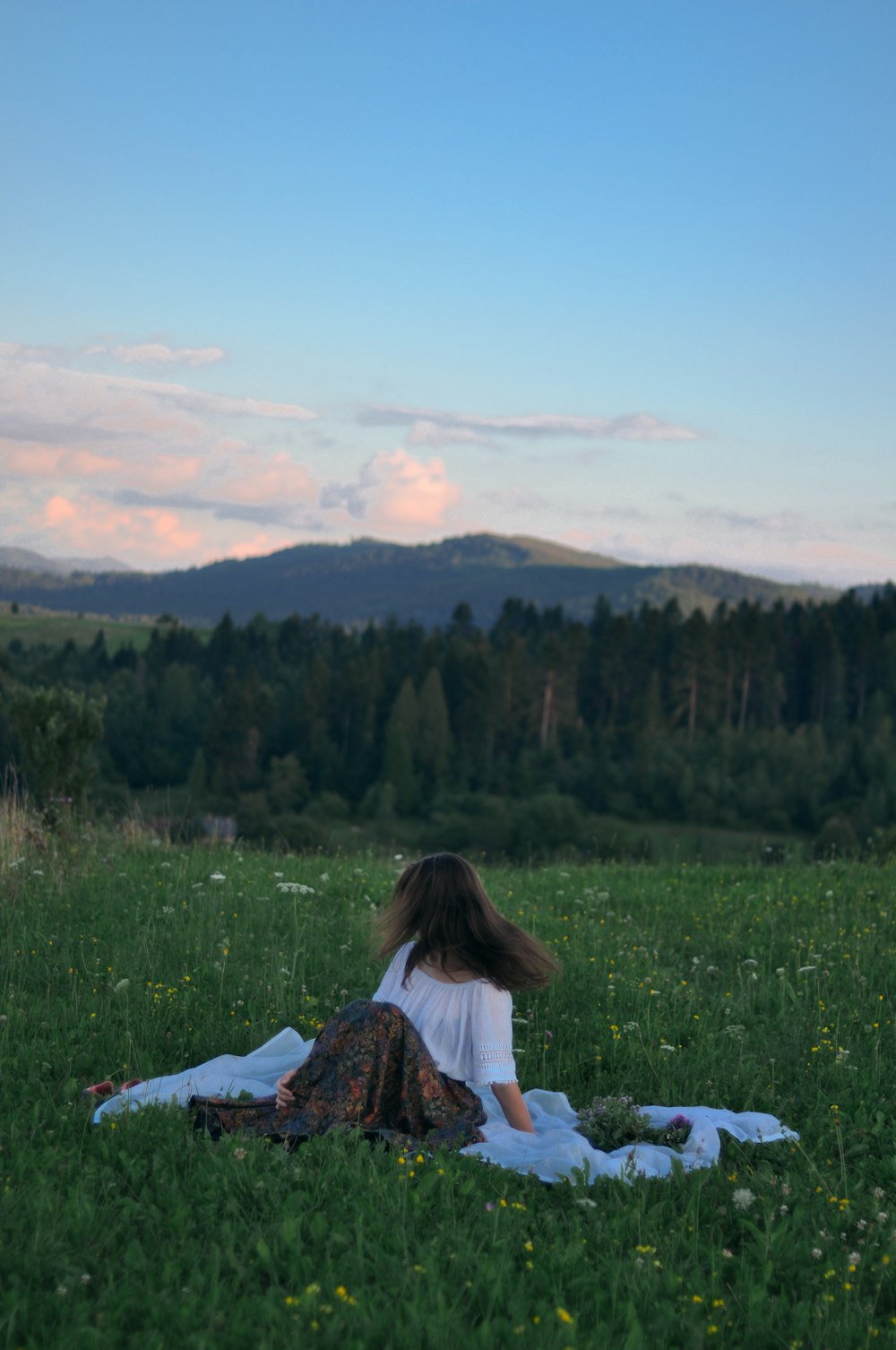 흰색과 빨간색 꽃 드레스를 입은 여자는 푸른 잔디밭에 파란색 직물에 앉아 있습니다.