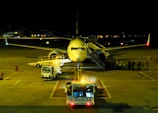 An airplane is parked on a dimly lit airport tarmac at night. Several maintenance vehicles with flashing lights are positioned around the aircraft. There is a line of passengers or crew members visible, boarding the plane via a set of mobile stairs.