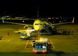 An airplane is parked on a dimly lit airport tarmac at night. Several maintenance vehicles with flashing lights are positioned around the aircraft. There is a line of passengers or crew members visible, boarding the plane via a set of mobile stairs.