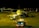 An airplane is parked on a dimly lit airport tarmac at night. Several maintenance vehicles with flashing lights are positioned around the aircraft. There is a line of passengers or crew members visible, boarding the plane via a set of mobile stairs.