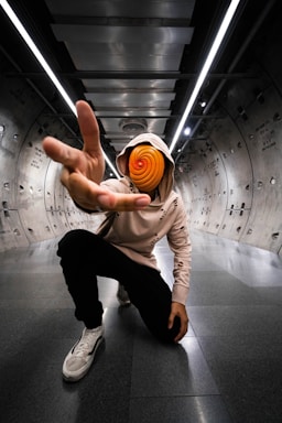 A person crouches in an industrial-style tunnel with metal walls and overhead lighting. They wear a pink hoodie with the hood up and black pants, extending their hand towards the camera. The person's face is obscured by a spiral, orange and red mask, creating a sense of mystery.