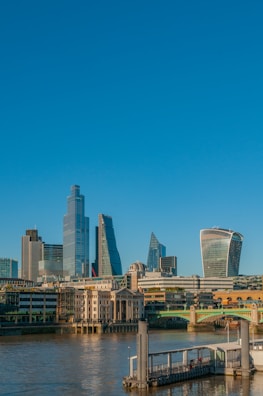 A modern city skyline with a mix of contemporary and iconic architecture. The buildings are highly diverse in design, featuring sleek glass structures and unique shapes. A river flows in the foreground with a dock and bridge, adding to the urban landscape.