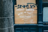 A wooden board is placed on a storefront with white paint reading 'Covent Garden We're in this together.' Above the text, there is blue graffiti. The scene includes part of a tree trunk on the left and a dark blue facade around the board.