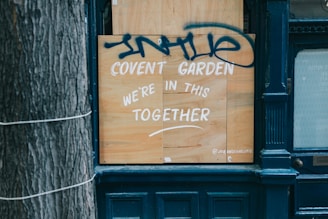 A wooden board is placed on a storefront with white paint reading 'Covent Garden We're in this together.' Above the text, there is blue graffiti. The scene includes part of a tree trunk on the left and a dark blue facade around the board.
