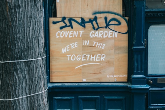 A wooden board is placed on a storefront with white paint reading 'Covent Garden We're in this together.' Above the text, there is blue graffiti. The scene includes part of a tree trunk on the left and a dark blue facade around the board.