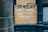 A wooden board is placed on a storefront with white paint reading 'Covent Garden We're in this together.' Above the text, there is blue graffiti. The scene includes part of a tree trunk on the left and a dark blue facade around the board.