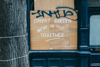 A wooden board is placed on a storefront with white paint reading 'Covent Garden We're in this together.' Above the text, there is blue graffiti. The scene includes part of a tree trunk on the left and a dark blue facade around the board.