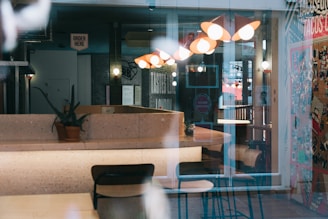 The image depicts an interior of a café or restaurant with modern lighting fixtures, tables, and chairs. A potted plant is placed on the countertop. There is a sign that reads 'Order Here', and a colorful wall filled with posters and graphics adds vibrancy to the setting. Reflections on glass surfaces subtly obscure the view.