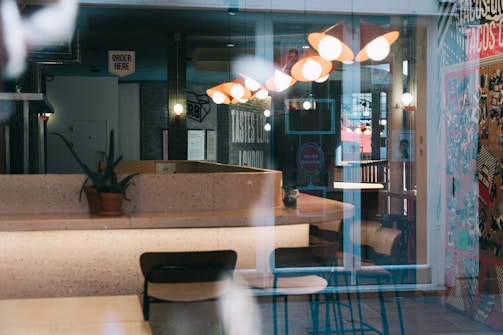 The image depicts an interior of a café or restaurant with modern lighting fixtures, tables, and chairs. A potted plant is placed on the countertop. There is a sign that reads 'Order Here', and a colorful wall filled with posters and graphics adds vibrancy to the setting. Reflections on glass surfaces subtly obscure the view.